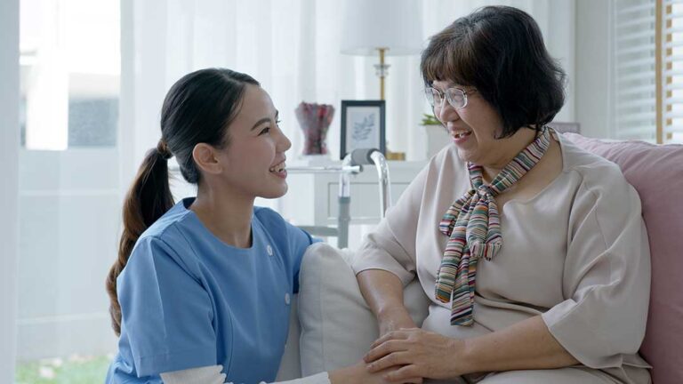 Two assisted living staff members helping an older adult with daily activities in a bright, residential common area.